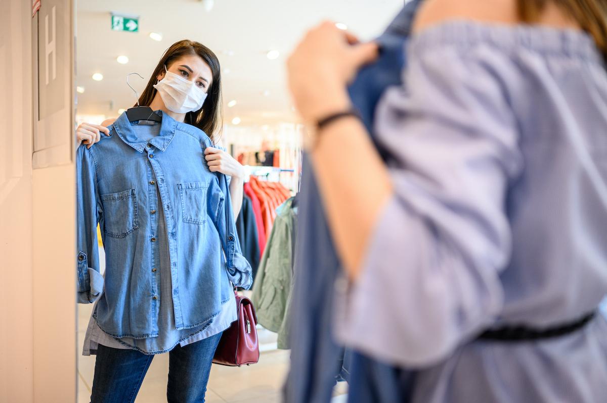 Woman wearing masks holds denim shirt up in front of her body in a store's mirror