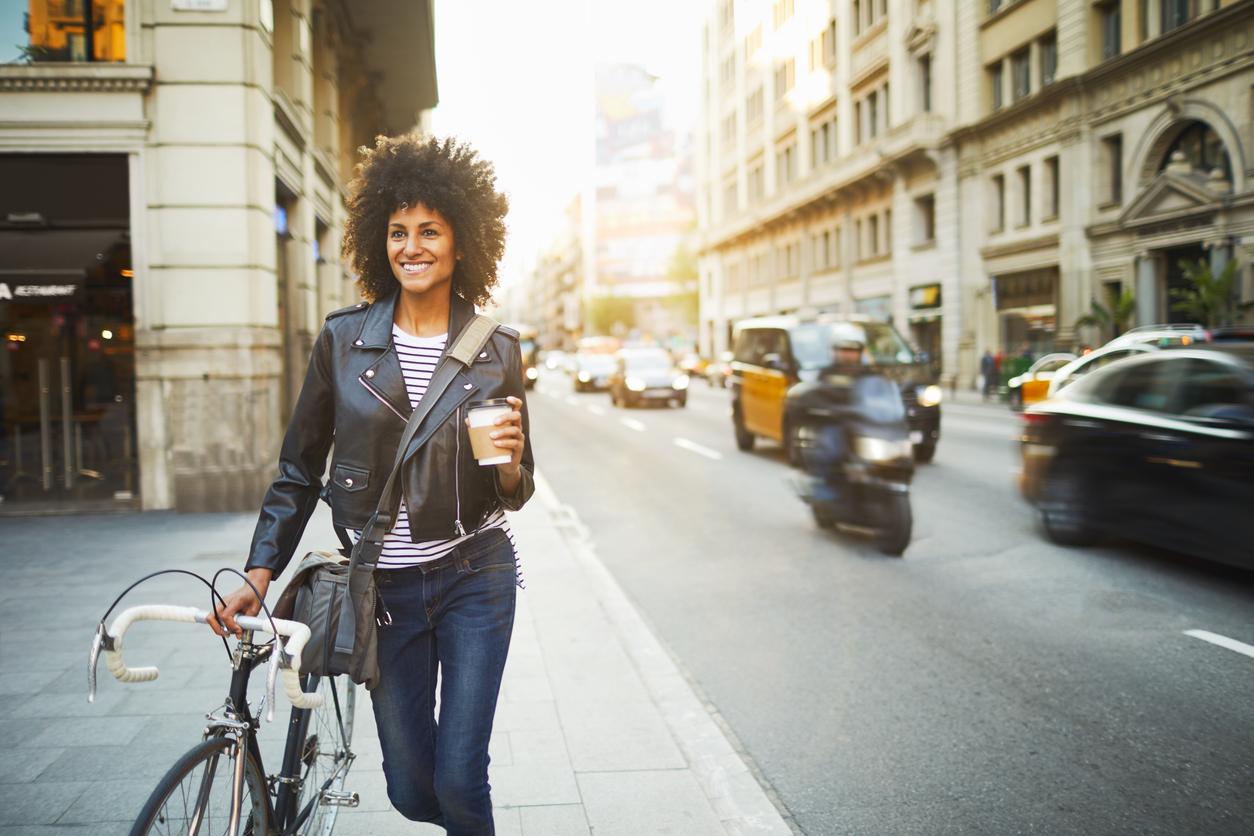 A photo of a smiling woman in a PU leather jacket walking with coffee in hand beside her bicycle on a city street.